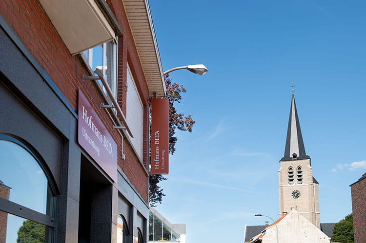 Gevel van een gebouw met het bord “Hofmans DELA Uitvaartzorg”, naast een straat met een kerktoren op de achtergrond onder een blauwe lucht.